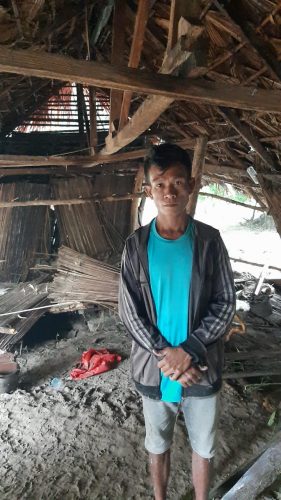 Paulus Mbura, warga desa Pukdale dengan latar belakang rumahnya yang porak-poranda diterjang banjir akibat meluapnya Kali Felakdale. (yandry/kupangterkini.com)
